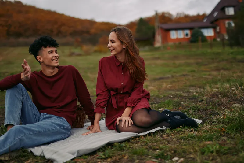 A young couple sitting on a blanket in a rural meadow, sharing a candid moment with genuine smiles and relaxed interaction that conveys authenticity and credibility in everyday life.