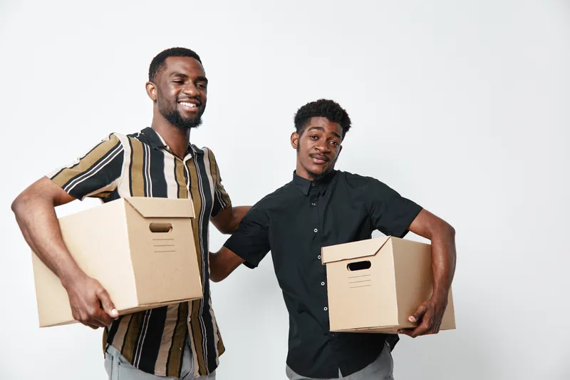 Two young African American men with dark skin holding cardboard boxes for moving or delivery, smiling and standing against white background in casual clothing for teamwork or relocation.