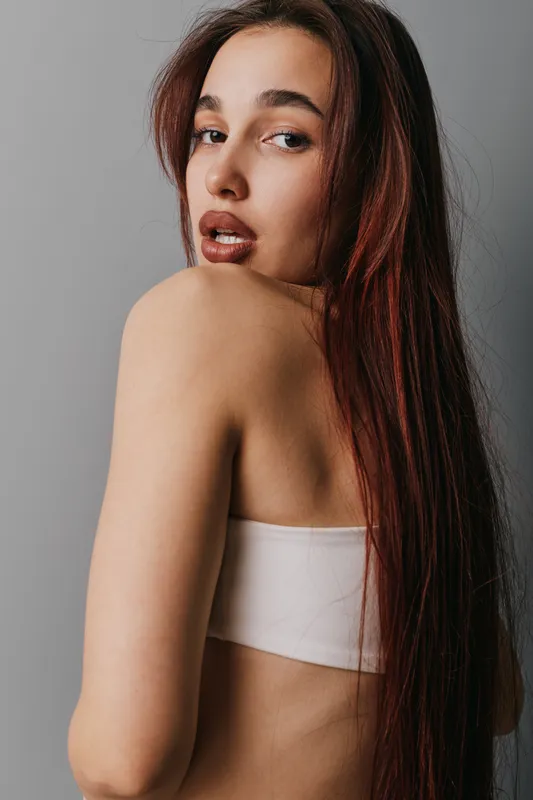 Young woman with long hair and natural makeup posing with bare shoulder looking over grey background. Beauty, fashion and portrait concept with casual style and confident expression.