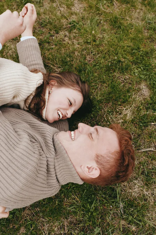 Candid couple lying on grass, smiling at each other in cozy sweaters. Close-up on faces, warm natural light, relaxed outdoor moment, lifestyle and affection.