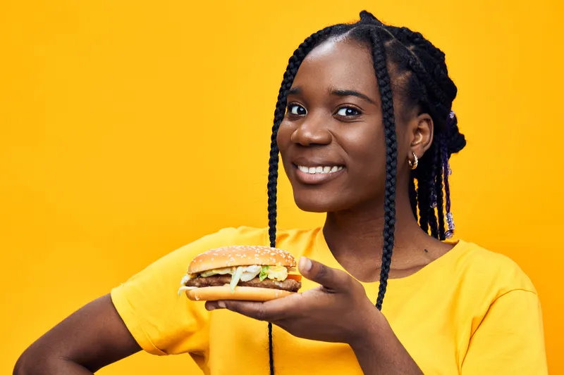 Young Black woman smiling while holding a delicious hamburger against a vibrant yellow background, showcasing joyful eating and fast food