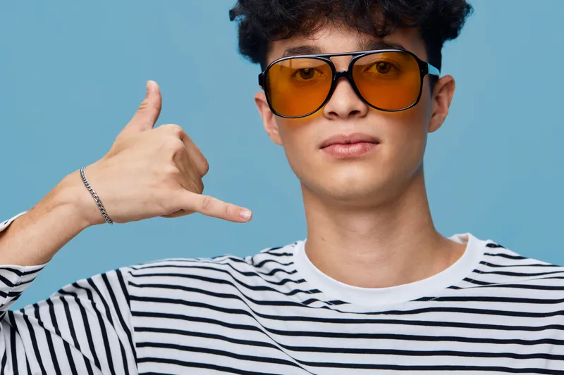 Young man wearing orange sunglasses and striped shirt making call me hand gesture against blue background, casual style with confident expression and modern look.