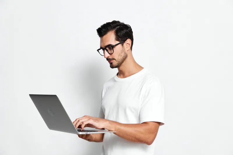 Full body focused thoughtful young man wearing a white t-shirt using a laptop while standing on a solid white background. Technology work concept.