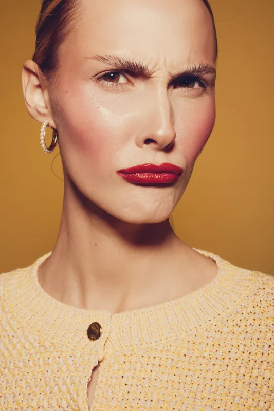 Close-up of a woman with red lipstick and hoop earrings frowning with a confused expression. She wears a textured beige sweater against a warm brown background. Concept of doubt and emotion.