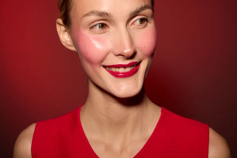 Smiling woman with bright red lipstick and flushed cheeks wears red sleeveless top against red background. Concept of beauty, makeup, fashion, and confident expression.