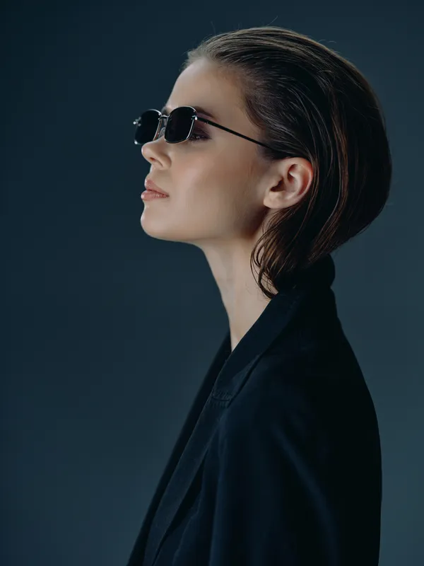 Profile of a stylish woman wearing dark sunglasses and a black blazer with a sleek hairstyle in a minimalistic dark studio setting.