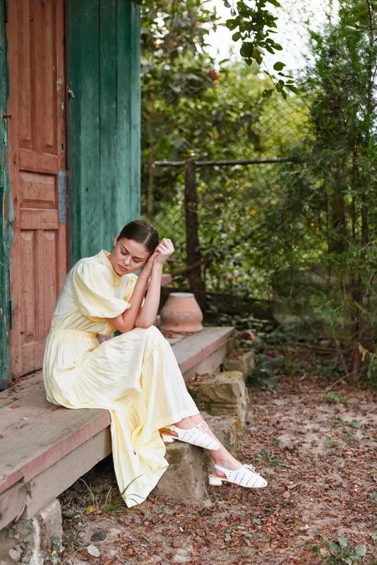 Woman in a yellow dress sits on wooden steps outdoors, looking thoughtful and calm in a lush garden setting with dappled sunlight and greenery, creating a serene contemplative mood.