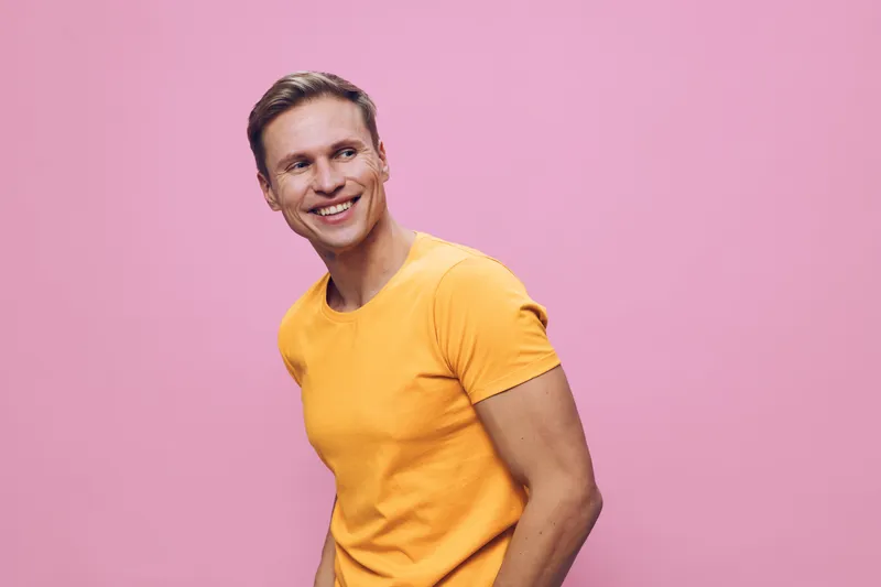 Smiling man wearing yellow t-shirt looking away against pink background, casual clothing, joyful expression, lifestyle portrait, vibrant colors, cheerful mood, studio shot.