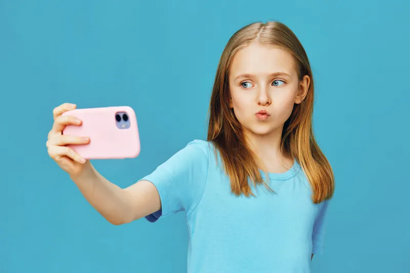 A young girl with long hair poses for a selfie against a vibrant blue background, showcasing a playful expression in a casual blue t shirt, perfect for social media moments