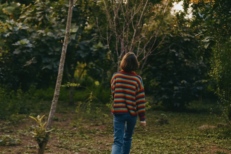 Person back in striped sweater walking through forest and nature, casual jeans and autumn foliage, solitary stroll on grassy path with relaxed posture and muted evening light