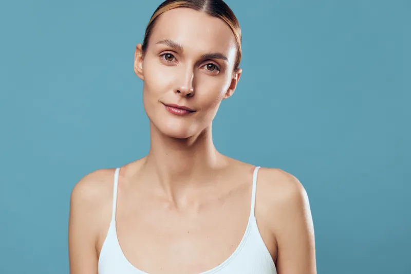 Portrait of a young woman with natural makeup and tied hair wearing a white tank top on a blue background. The concept of beauty, wellness, and minimalism with soft lighting.