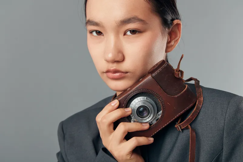Young woman holding vintage camera close to face in studio, looking at camera. Portrait of female photographer with retro analog camera in leather case against grey background.