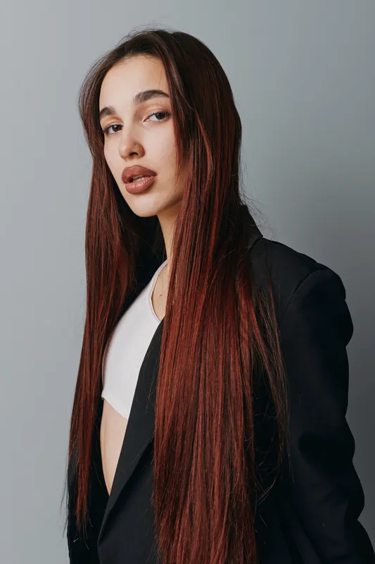 Young woman with long straight hair wearing black blazer and white top posing against gray background with serious expression and natural makeup for fashion portrait.