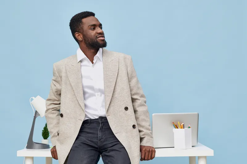 Professional stylish man wearing beige blazer and white shirt standing confidently in modern office with light blue background, workspace decor, and laptop, portraying success, confidence, and busines