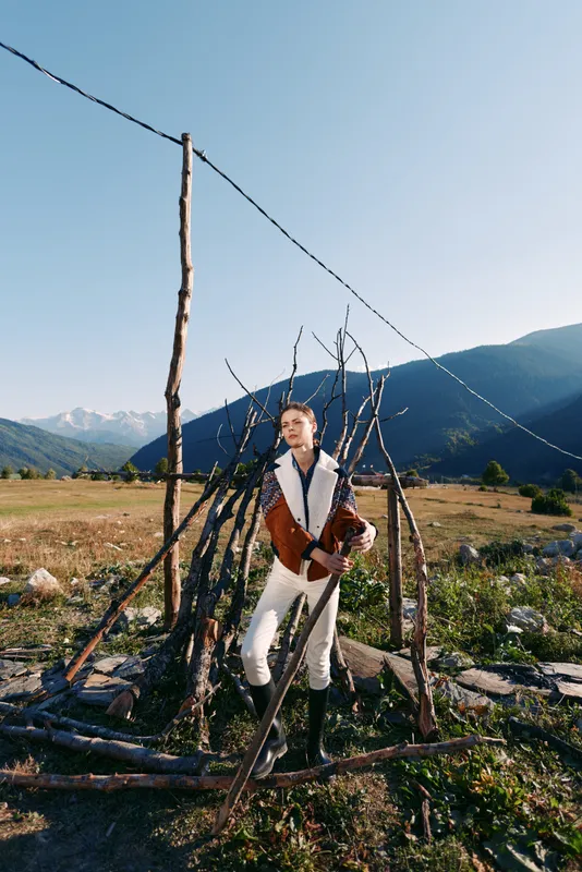 Man musician with bagpipes stands in a mountain meadow by a rustic fence, wearing traditional folk costume and boots, rural landscape with wooden stakes and distant snowcapped peaks.