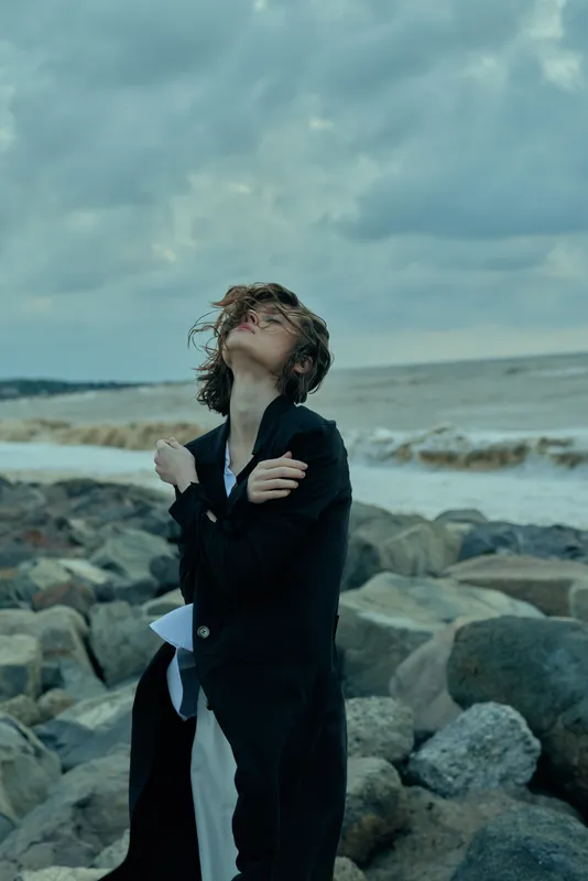 Windy shore, ocean rocks, coat moody sky weather, wind solitary figure, coastal scene, fashion outdoors, landscape sea breeze rugged coast, waves hair windblown clouds atmospheric dramatic light mood