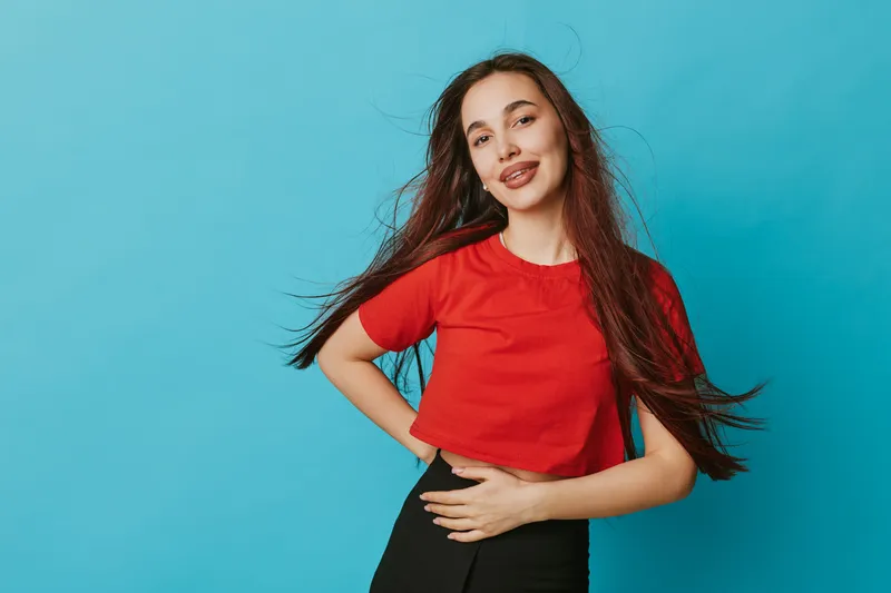 Young woman with long hair wearing red shirt and black pants posing with hand on waist and smiling against blue background in studio. Casual style and confident expression.