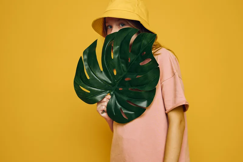 Girl wearing yellow hat and pink shirt hides half face behind large green leaf on yellow background. Concept of mystery and fashion portrait.