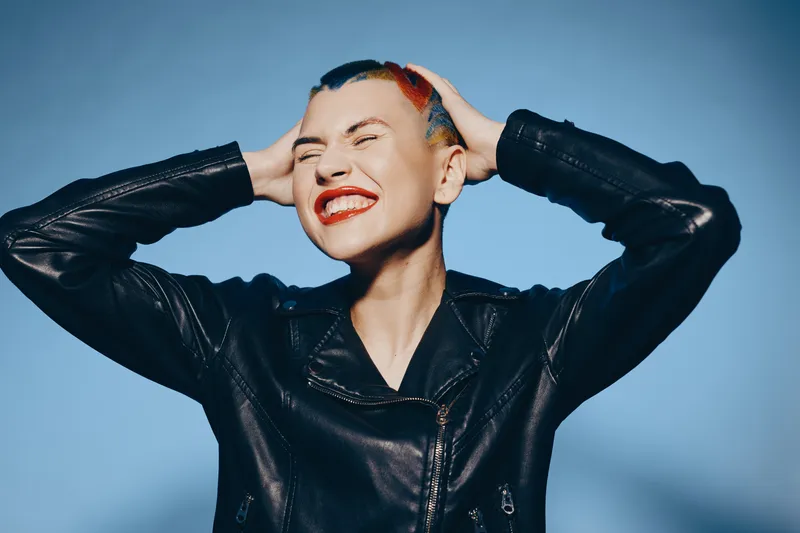 smiling woman with short colorful hair wearing black leather jacket, joyful expression with closed eyes, bright red lipstick, blue background studio portrait