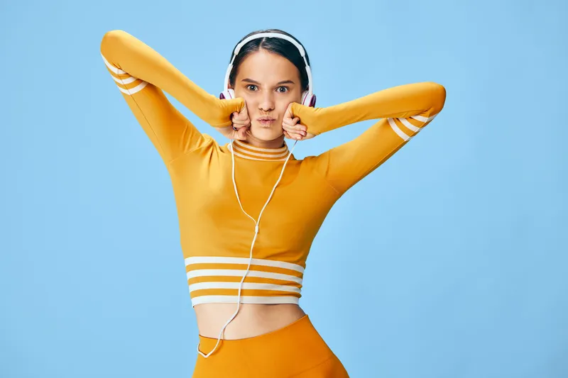 Woman fitness portrait in a yellow athletic crop top and high waisted leggings wearing headphones on a blue studio background energetic sporty woman fitness concept with playful pose