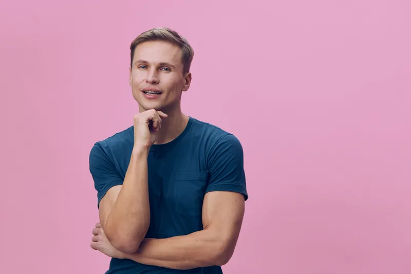 Confident young man with blonde hair, wearing casual blue t-shirt, posing thoughtfully with hand on chin against pink background, studio shot, portrait, people lifestyle concept, modern fashion.