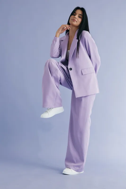 Stylish woman in lavender pantsuit posing confidently against a soft purple background, showcasing contemporary fashion and casual white sneakers in a studio setting.