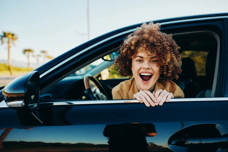 Joyful woman leans from car window, smiles widely, celebrating a carefree lifestyle with retro film color mood, sunny street scene, candid moment, vibrant energy with optimistic vibe