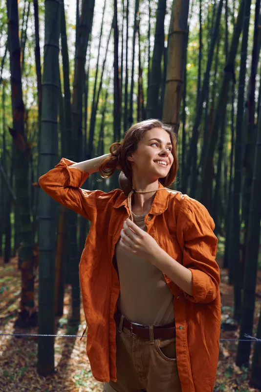 Woman in bamboo forest portrait, nature scene with warm smile, casual outfit and relaxed pose outdoors, sunlight filtering through tall trunks creating peaceful mood.