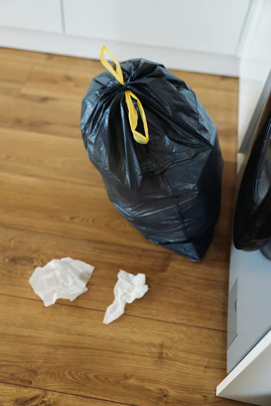 Trash garbage bag kitchen waste floor scene with a black tied plastic sack next to cabinets and crumpled tissues on hardwood, domestic disposal and messy cleaning situation.