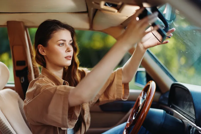 Young woman adjusting her rearview mirror in a vintage car with a relaxed expression, embodying a sense of freedom and adventure during summer.