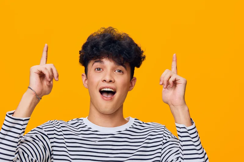 young man with curly hair wearing striped shirt excitedly pointing up against bright orange background, expressing surprise and joy in casual style