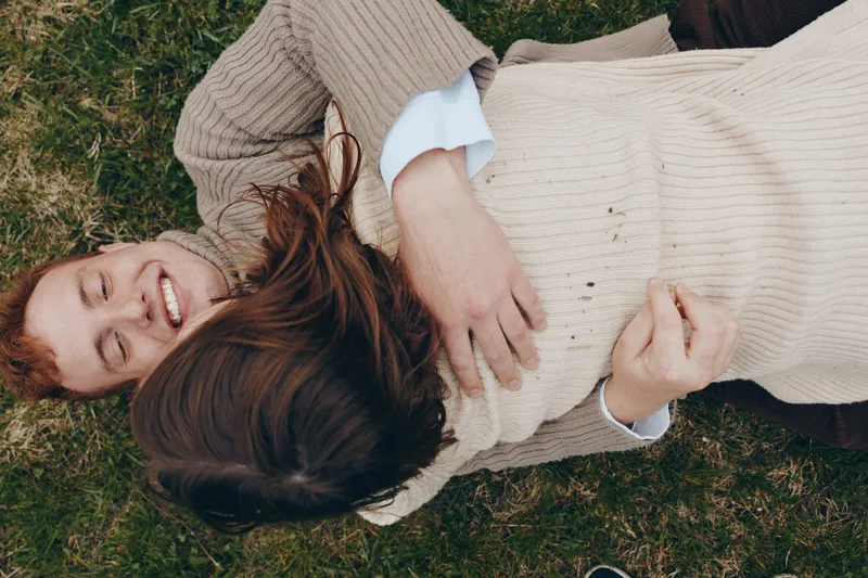 Pregnant woman lying on grass with partner, smiling, relaxed. Casual sweater, soft natural light, warm mood. Lifestyle portrayal of family, wellbeing, and anticipation of motherhood in an outdoor