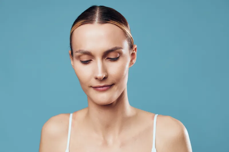 Calm young woman with closed eyes and smooth skin poses against blue background, wearing white tank top, showing peaceful and relaxed facial expression.