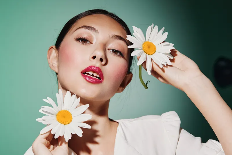 Fashionable young woman with glossy red lips and pastel makeup holding daisies near her face on soft green background flash light highlights gloss effect.