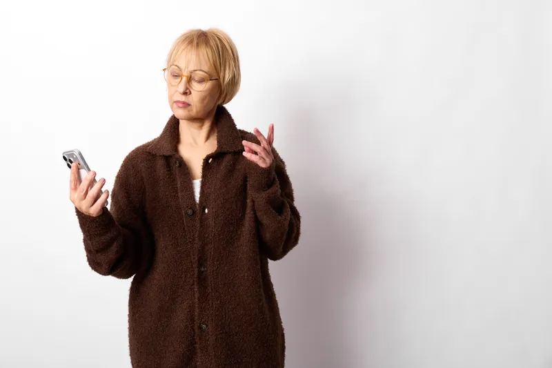 Woman with smartphone shows stress and anxiety, frustration and distress, she touches hair and gestures while standing on a clean white background, symbol of technology overload, mental health