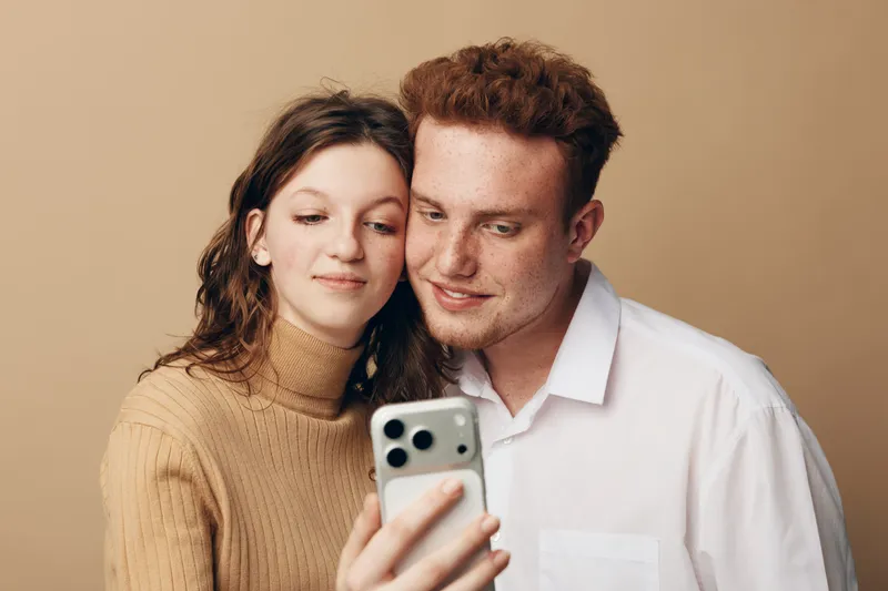Young couple with red hair and brown hair takes selfie on smartphone. They smile gently and look at screen in casual clothes on beige background. Concept of friendship and technology.