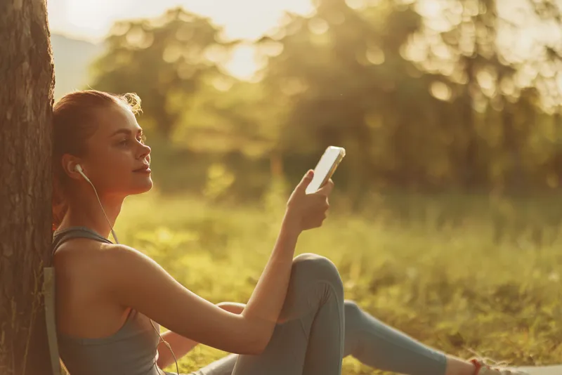 Young woman enjoying music on her smartphone in a sunlit park, evoking relaxation and tranquility through a serene outdoor setting