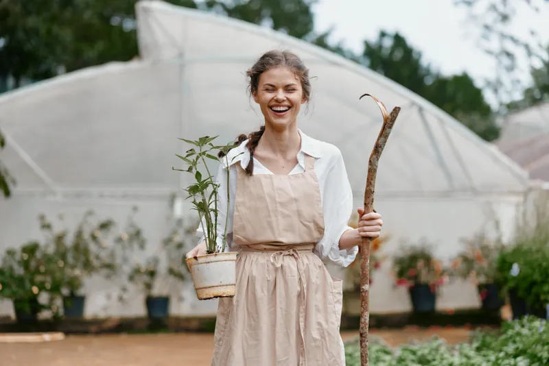 Happy young woman holding a potted plant and a stick in a garden, wearing an apron, with a greenhouse in the background, showcasing passion for gardening and nature