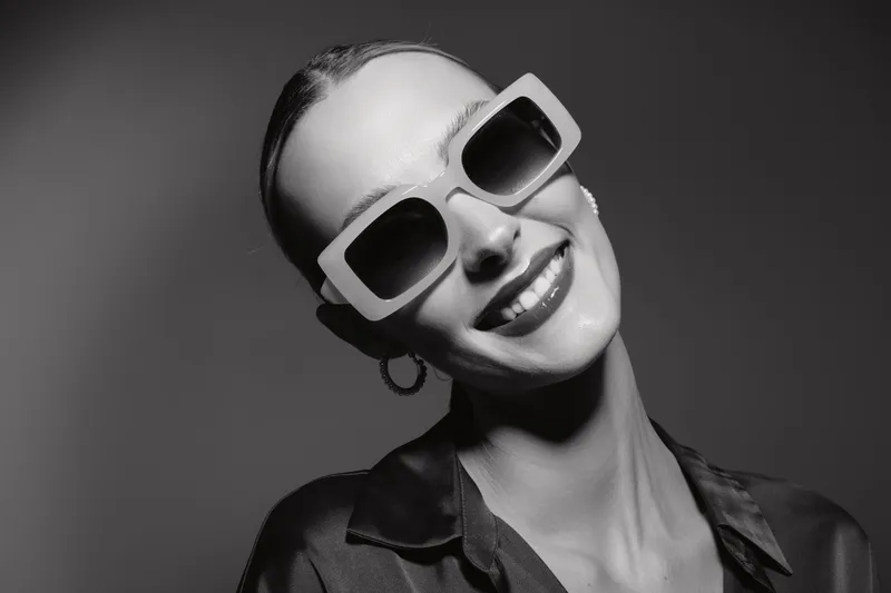 Smiling Caucasian woman with pale skin wears large white sunglasses and hoop earrings. She poses confidently with a bright smile against a dark background in black and white.