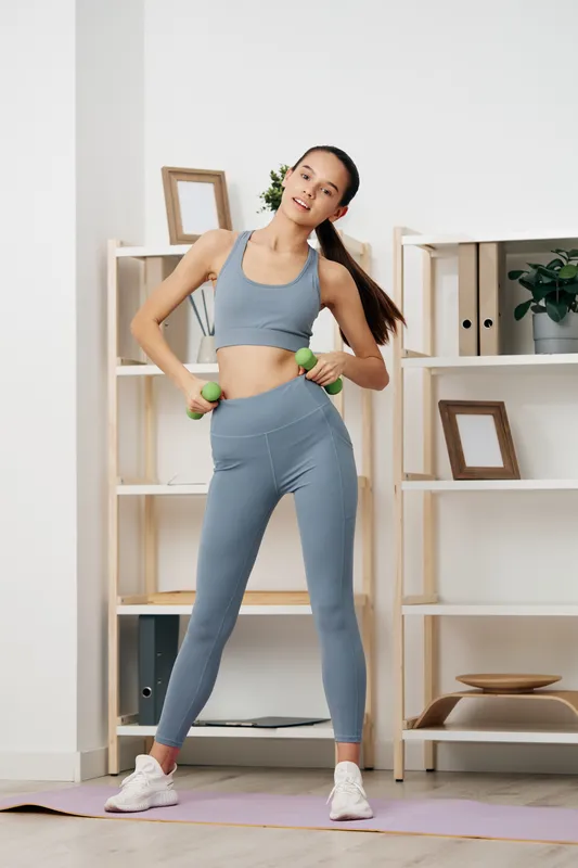 Fitness woman in athletic wear demonstrates a safe home workout with dumbbells, performing standing moves in a bright living space for motivation.