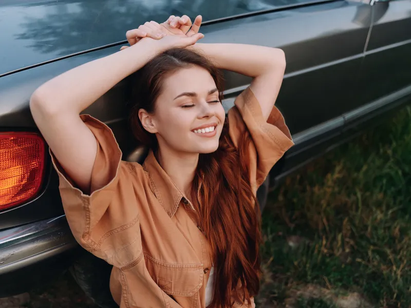 Young woman with long hair in a casual brown shirt, smiling happily against a vintage car. Sunlit outdoor scene conveying joy and relaxation.