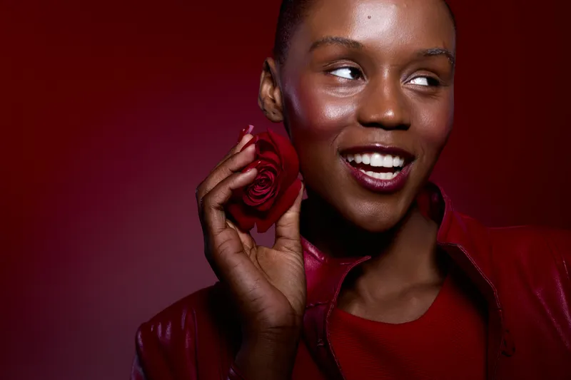 Smiling dark skinned African woman with short hair holds red rose near face. She wears red jacket and dress against dark red background. Concept of beauty, happiness, and floral elegance.