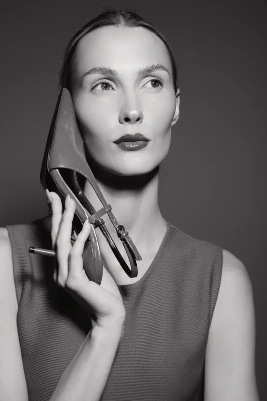 Caucasian white woman with fair skin holds high heel shoe near face looking away. Concept for fashion, elegance, and modern style in black and white portrait.