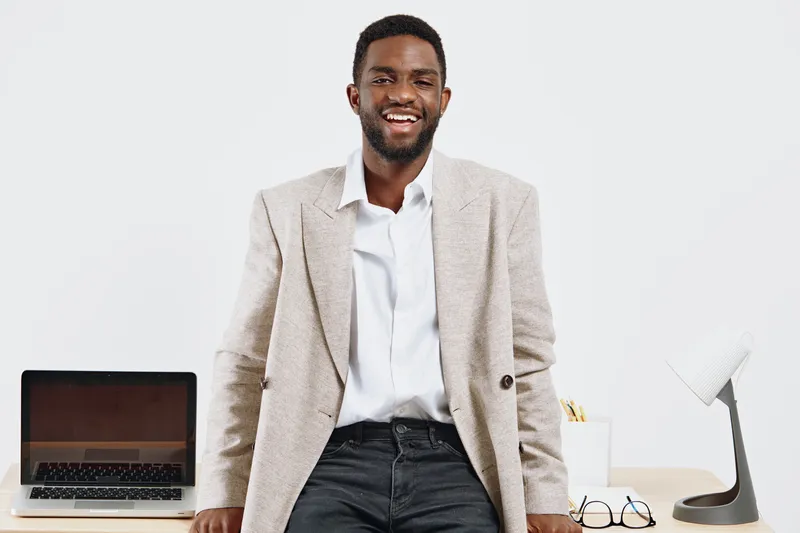 Confident smiling man of African descent with dark skin, wearing beige blazer and white shirt, standing in modern office with laptop, glasses, and desk accessories, professional business attire, cheer