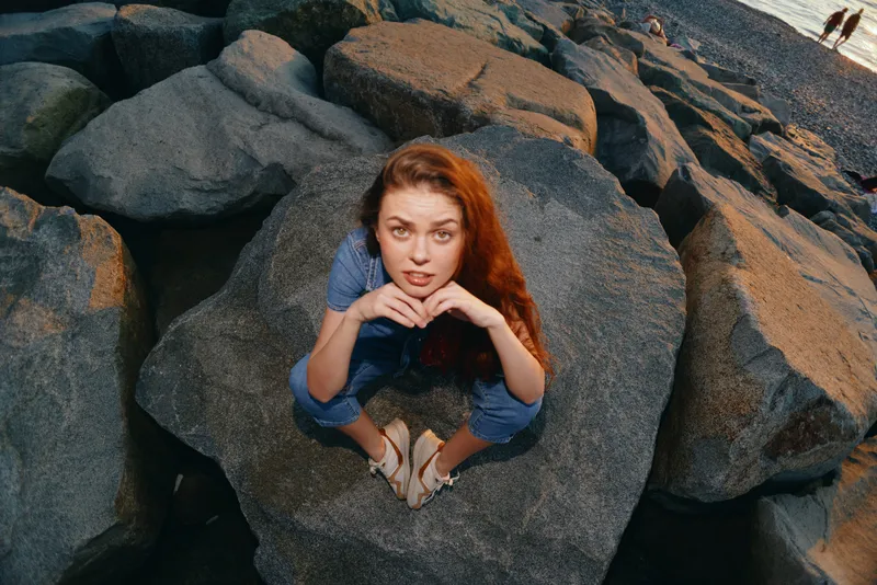 young woman sitting on large rocks by the sea wearing casual blue outfit and sandals during golden hour with thoughtful expression