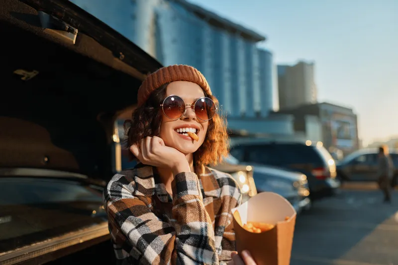 Young woman with a bright smile leaning on a car trunk eating fries, wearing sunglasses in an urban sunset. Candid lifestyle shot emphasizing authenticity, golden hour glow, mindful living and