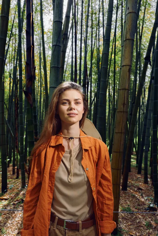 Woman bamboo forest portrait, standing outdoors in nature wearing orange jacket and beige blouse. Young female with confident expression among vertical trees in a serene woodland setting.
