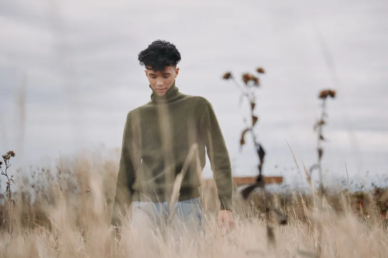 A young person stands in tall dry grass wearing a green sweater, captured in soft natural light to convey quiet authenticity and realism, candid posture, gentle expression and introspective mood.