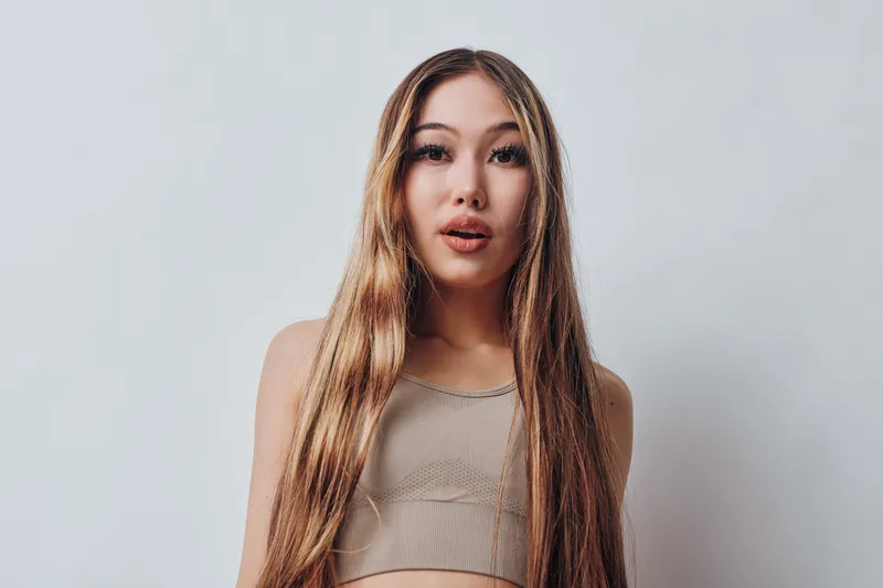 Young woman with long straight hair and natural makeup wearing a beige top standing against plain light background in studio portrait.