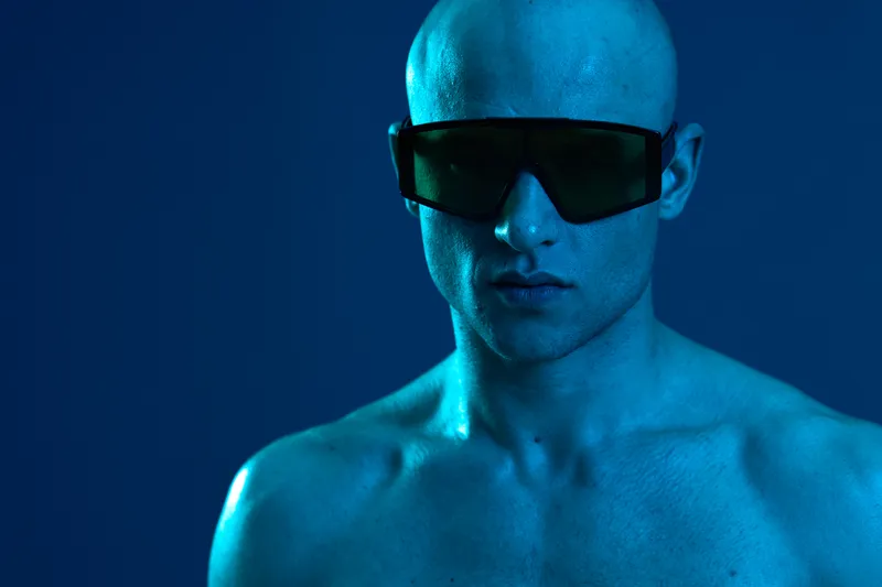 Man with shaved head wearing sunglasses, showcasing a strong expression against a vibrant blue background. The image has a dynamic and modern vibe, perfect for fashion themes.